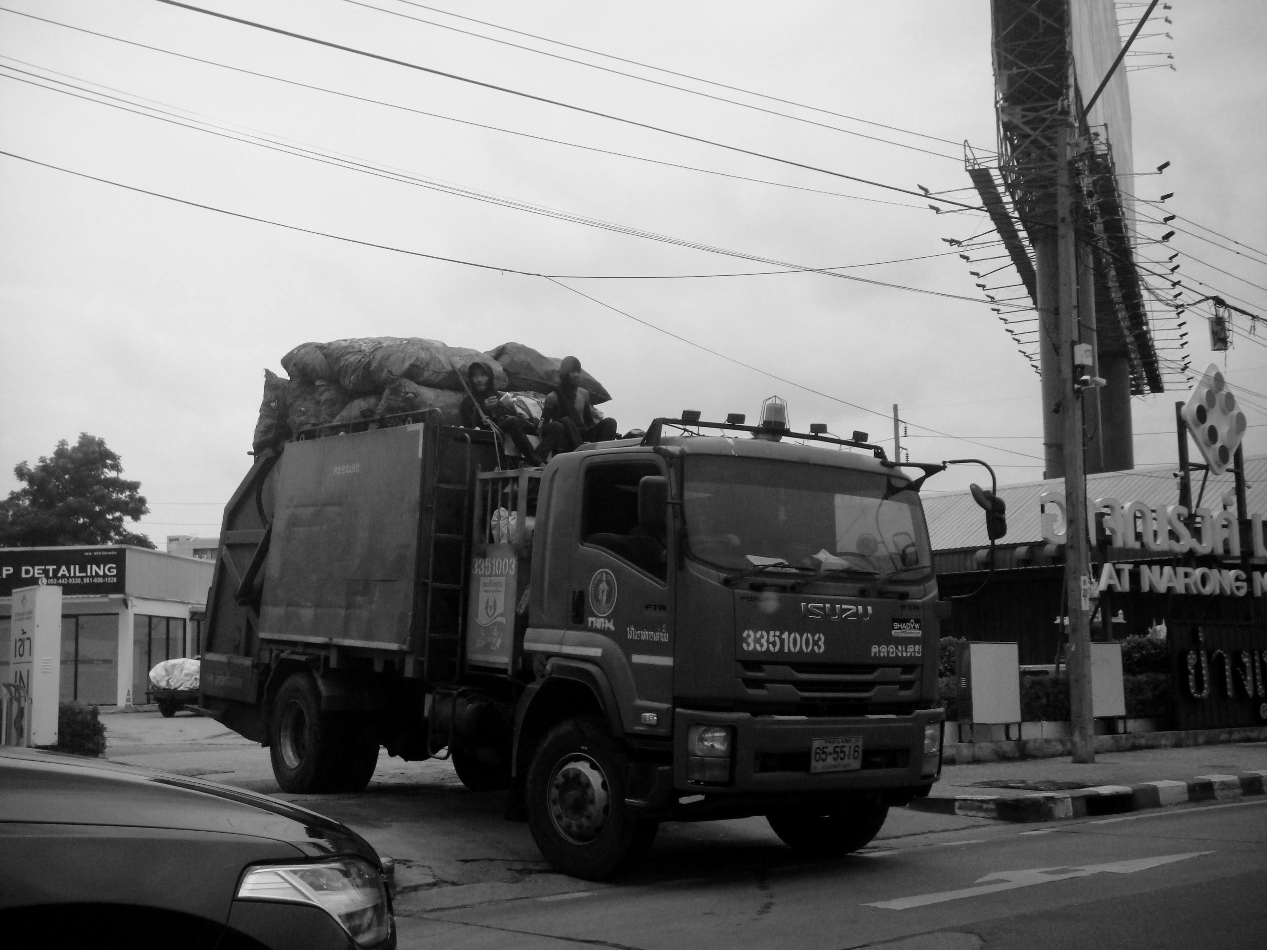 Black-and-white street photograph of a large truck carrying a covered load, with two people seated behind the cab and overhead wires crossing the sky.