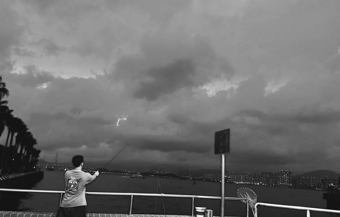 Black-and-white image of a person fishing from a waterfront railing, with palm trees, a distant city skyline, and heavy clouds over the water.