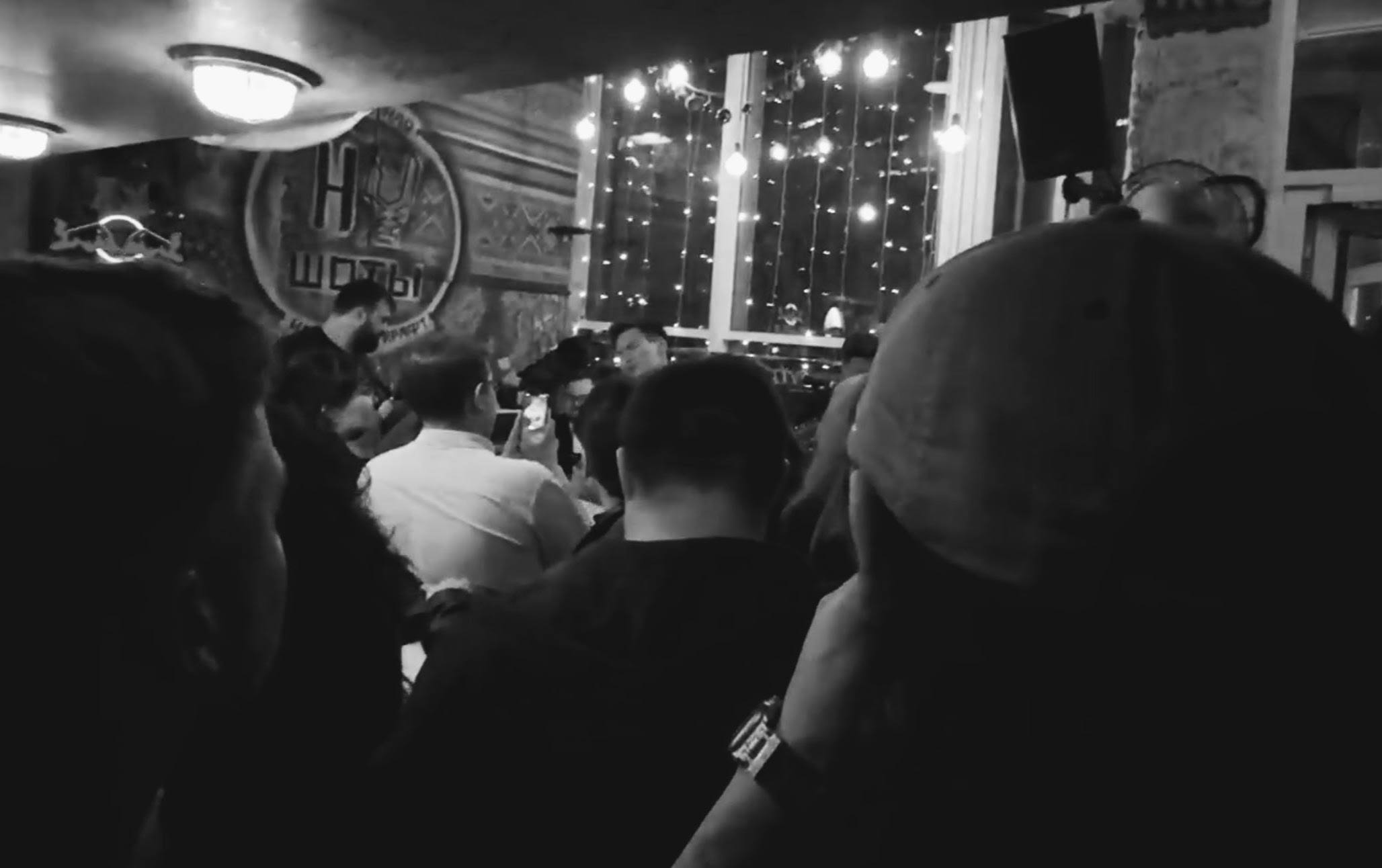 Black-and-white image of a band performing in a crowded bar, seen over the audience with string lights hanging behind the musicians.