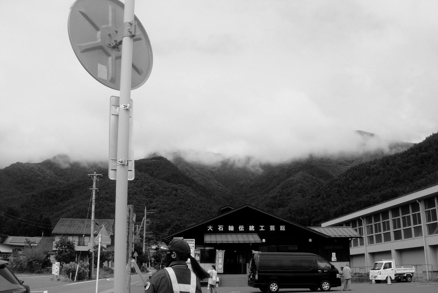 Black-and-white photograph of fog hanging over wooded mountains near Mount Fuji, with roadside buildings, vehicles, and a person in the foreground.