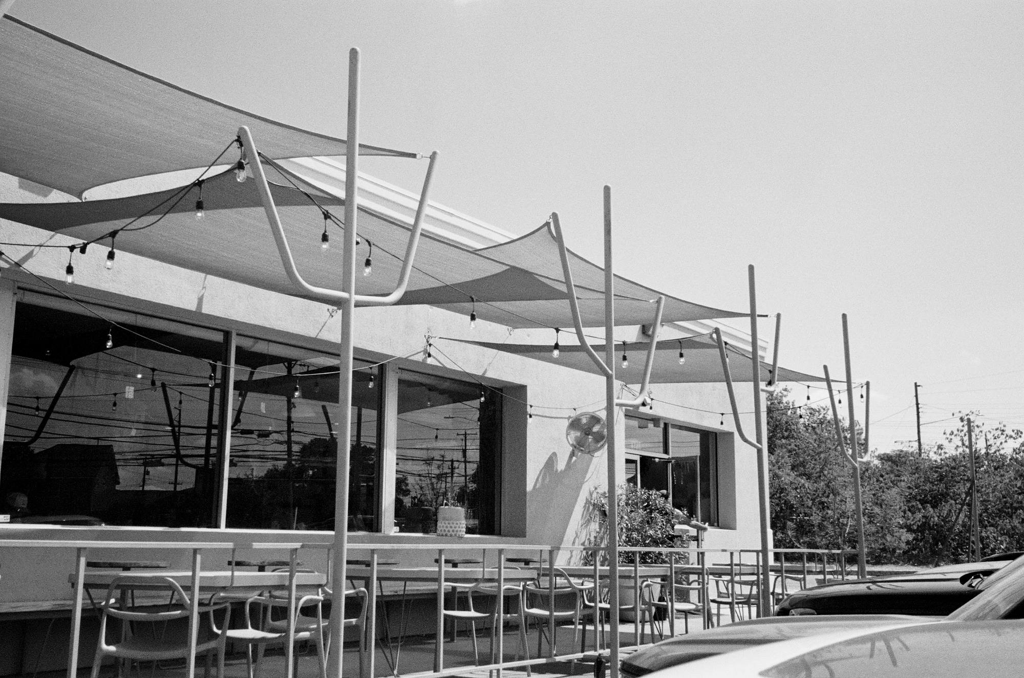 Shade sails and string lights over an empty patio, bare chairs and a long counter, midday.